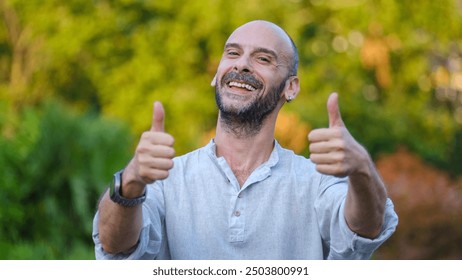 A middle-aged handsome businessman looking at the camera in a tree-lined park on a summer day, giving a thumbs-up to show approval - Powered by Shutterstock