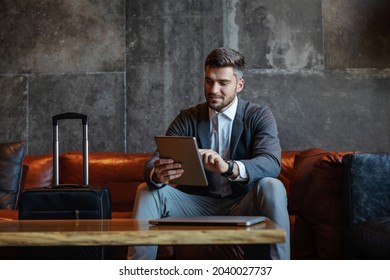 Middle-aged handsome businessman sitting on a couch in a hotel lobby and check his online social networks on a digital tablet. Next to him is luggage. Business trip, travel, technologies - Powered by Shutterstock