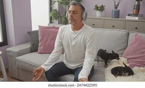 Middle-aged hispanic man sits in a living room with two chihuahuas, wearing headphones and meditating on a couch surrounded by modern indoor decor. - Powered by Shutterstock