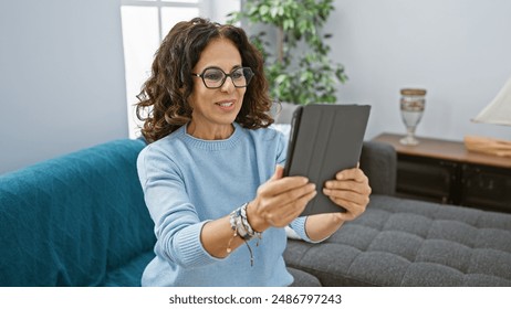 Middle-aged hispanic woman with curly hair using a tablet in a cozy living room at home, exhibiting technology and leisure. - Powered by Shutterstock