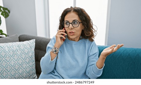 A middle-aged hispanic woman with curly hair talks on the phone in the living room of her apartment. - Powered by Shutterstock