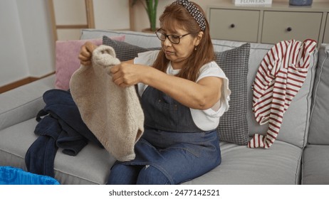 A middle-aged hispanic woman is folding knitted clothes in her living room, surrounded by laundry and sitting on a cozy sofa. - Powered by Shutterstock