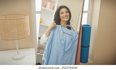 A middle-aged hispanic woman holds a blue shirt, smiling in a well-lit room with a yoga mat visible. - Powered by Shutterstock