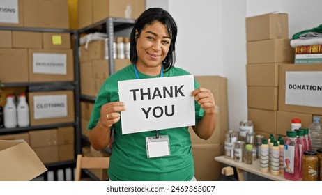Middle-aged hispanic woman holds 'thank you' sign in a donation center with boxes and supplies. - Powered by Shutterstock - Get 15% off with code: PIKWIZARD15