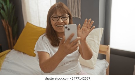 Middle-aged hispanic woman making a video call on her smartphone while sitting on a bed in her cozy bedroom. - Powered by Shutterstock