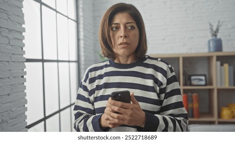 A middle-aged hispanic woman in a striped sweater looks concerned while holding a smartphone in a modern living room. - Powered by Shutterstock