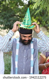 A Middle-aged Indian Man With A Turban On His Head Trying To Wear A Paper Hat For A Birthday