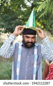 A Middle-aged Indian Man With A Turban On His Head Trying To Wear A Paper Hat For A Birthday