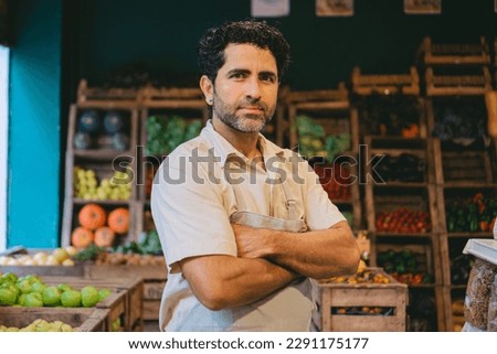 Middle-aged Latin man looking seriously at the camera with his arms crossed in a greengrocer's shop.