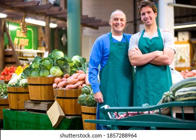 Middle-aged male grocer standing in front of his shop with an arm around his young son and smiling at the camera