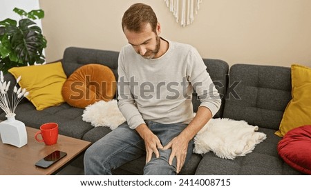 A middle-aged man with a beard grimaces in pain while holding his leg in a modern living room.