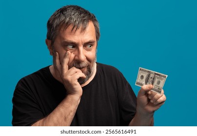 A middle-aged man with a beard holds a hundred dollar bill while looking contemplative. His hand rests on his face, suggesting he is pondering an important decision or idea. - Powered by Shutterstock