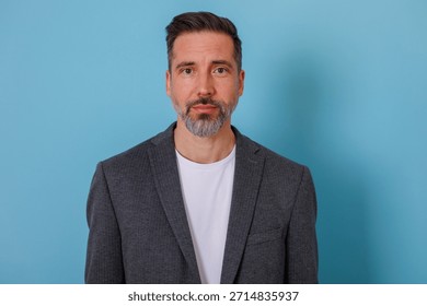 Middle-aged man with beard posing confidently in professional blazer in front of a blue background, looking at camera