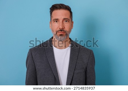 Middle-aged man with beard posing confidently in professional blazer in front of a blue background, looking at camera