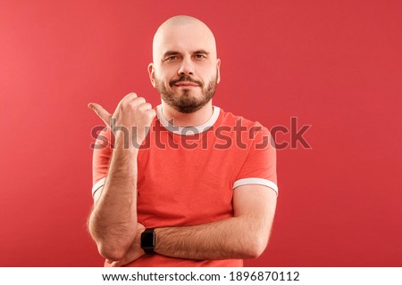 A middle-aged man with a beard in a red T-shirt on a red background joyfully points to the right. Isolated