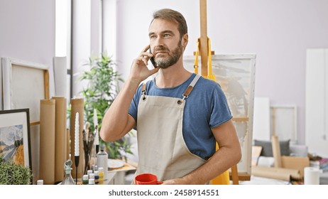 A middle-aged man with a beard stands in a studio, phone in hand, surrounded by art supplies and paintings. - Powered by Shutterstock