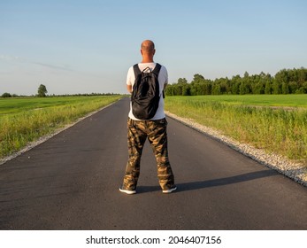 A Middle-aged Man With A Black Backpack Is Standing On The Asphalt Road With His Back To The Camera. The Man Folded His Arms Across His Chest. The Concept Of Travel And New Discoveries