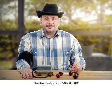 Middle-aged Man In Black Cowboy Hat Takes His Blood Pressure And Writes Down Result. Bearded Man With Cuff Around His Right Arm Sits At Wooden Desk Looking Directly At Camera