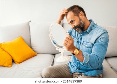 Middle-aged man in a casual denim shirt looking at hair loss in a handheld mirror while sitting on a sofa. - Powered by Shutterstock