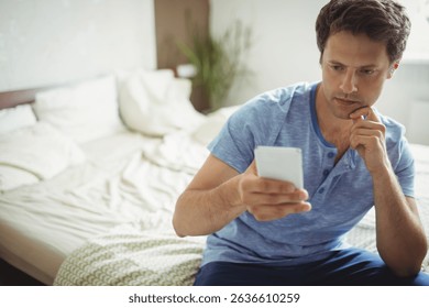 Middle-aged man checking smartphone while resting chin on hand on bed with plant, copy space. Casual, relaxation, home interior, modern, daily routine, cozy, peaceful