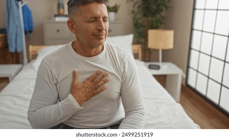 Middle-aged man clutching chest in distress sitting on bed in a cozy bedroom with soft lighting and comfortable furnishings. - Powered by Shutterstock