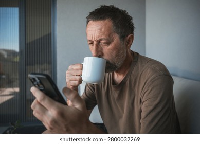 Middle-aged man drinking coffee while checking smartphone on balcony at home in morning light, selective focus