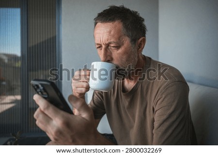 Middle-aged man drinking coffee while checking smartphone on balcony at home in morning light, selective focus