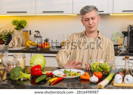 Middle-aged man eats vegetable salad with frustration during strict weight loss routine in kitchen. Young adult guy shows dislike for healthy food while testing willpower and nutrition habits at home.