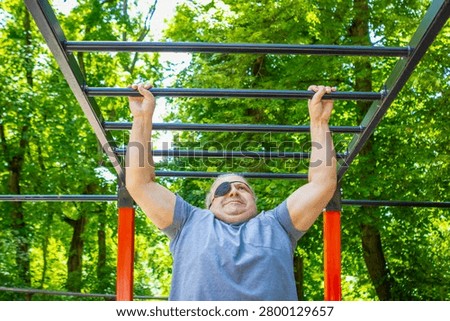 Middle-aged man with eyepatch performs pull-up on monkey bars in sunny park, showing strength and determination.