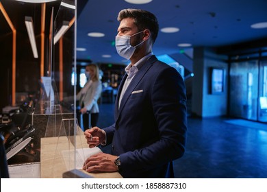 A Middle-aged Man In Formal Wear With A Face Mask Standing On The Reception Of A Fancy Hotel And Checking In.