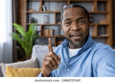 Middle-aged man gesturing with finger while talking during video call at home. Engaging and communicative interaction suggesting explanation or teaching moment. Comfortable living space