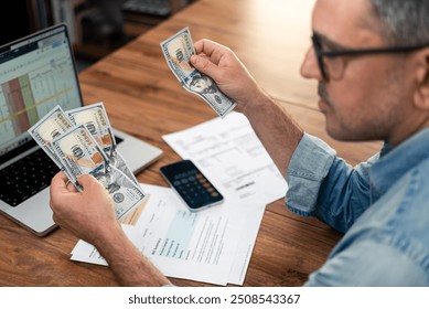 Middle-aged man with glasses reviewing dollar bills while working on household expenses at a desk. Financial planning utility bills and a laptop. Home finance and payment management concept.  - Powered by Shutterstock