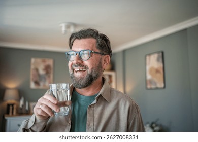 Middle-aged man with glasses smiling while holding a glass of water in a cozy home setting, conveying a sense of relaxation and contentment