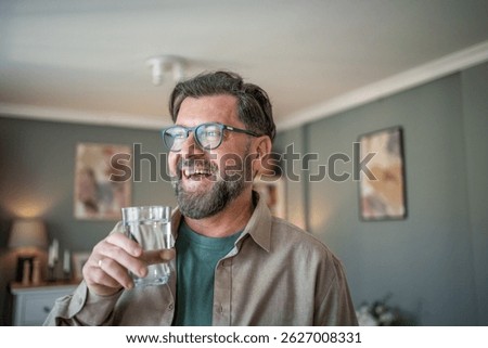 Middle-aged man with glasses smiling while holding a glass of water in a cozy home setting, conveying a sense of relaxation and contentment