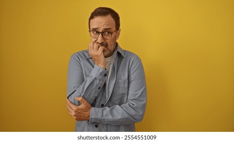 Middle-aged man with glasses standing against a vibrant yellow background appears thoughtful and concerned, dressed in casual attire with a hand to his mouth. - Powered by Shutterstock