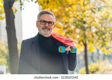 Middle-aged Man With Grey Beard And Hair Wearing Glasses And Blazer Smiling Looking Into Camera Holding A Colorful Skateboard On His Shoulder. Fun Grandpa. Horizontal Outdoor Shot. High Quality Photo