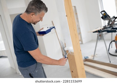 Middle-aged man with grey hair painting wooden beams in attic apartment, construction site with white paint
