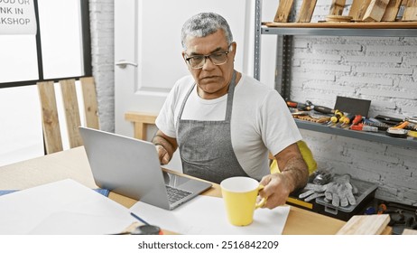 A middle-aged man with grey hair reviews plans on a laptop in a carpentry workshop while holding a coffee cup. - Powered by Shutterstock