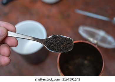 Middle-aged Man Holding Spoonful Of Dry Tea. Crushed Black Tea Leaves In Spoon Over Red Table. Cup And Container Are Blurred On Background. Healthy Lifestyle. First-person View. Front Focus. Indoor.