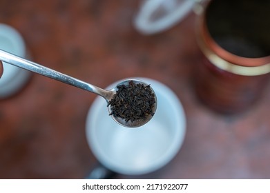 Middle-aged Man Holding Spoonful Of Dry Tea. Crushed Black Tea Leaves In Spoon Over Red Table. Cup And Container Are Blurred On Background. Healthy Lifestyle. First-person View. Front Focus. Indoor.