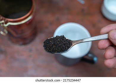 Middle-aged Man Holding Spoonful Of Dry Tea. Crushed Black Tea Leaves In Spoon Over Red Table. Cup And Container Are Blurred On Background. Healthy Lifestyle. First-person View. Front Focus. Indoor.