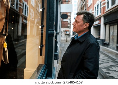 Middle-aged man in a jacket observing a store's window display with a reflection of buildings on an urban city street, contemplating consumerism and aspiration