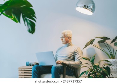 Middle-aged man looking aside while working on laptop while sitting in armchair at home. Confident, experienced senior male professional. Small business entrepreneur manage business at home.