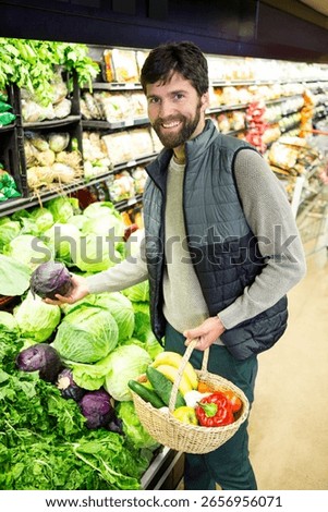 Middle-aged man picking purple cabbage and holding basket with bananas in supermarket produce aisle. Freshvegetables, grocery shopping, localmarket, farmtotable, healthyfood, organicproduce,
