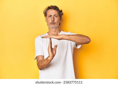 Middle-aged man posing on a yellow backdrop showing a timeout gesture.