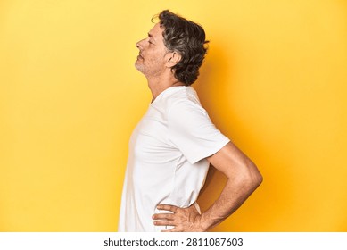 Middle-aged man posing on a yellow backdrop suffering a back pain.