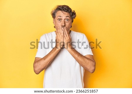 Middle-aged man posing on a yellow backdrop shocked, covering mouth with hands, anxious to discover something new.