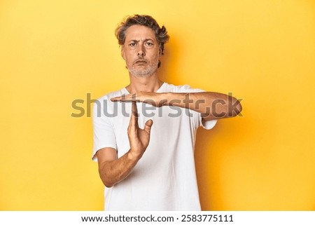 Middle-aged man posing on a yellow backdrop showing a timeout gesture.