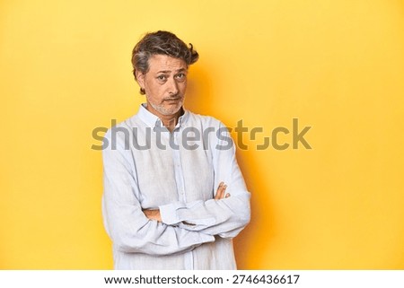 Middle-aged man posing on a yellow backdrop suspicious, uncertain, examining you.