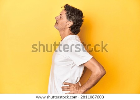Middle-aged man posing on a yellow backdrop suffering a back pain.